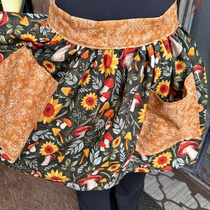 K&ouml;nnte beinhalten: Ein farbenfrohe Sch&uuml;rze mit dunkelgr&uuml;nem Hintergrund, verziert mit Sonnenblumen, Pilzen und Bl&auml;ttern. Die Sch&uuml;rze hat zwei Taschen und einen Bund mit kontrastierendem orangefarbenem Blumenmuster. F&uuml;r K&uuml;che oder Bastelarbeiten.