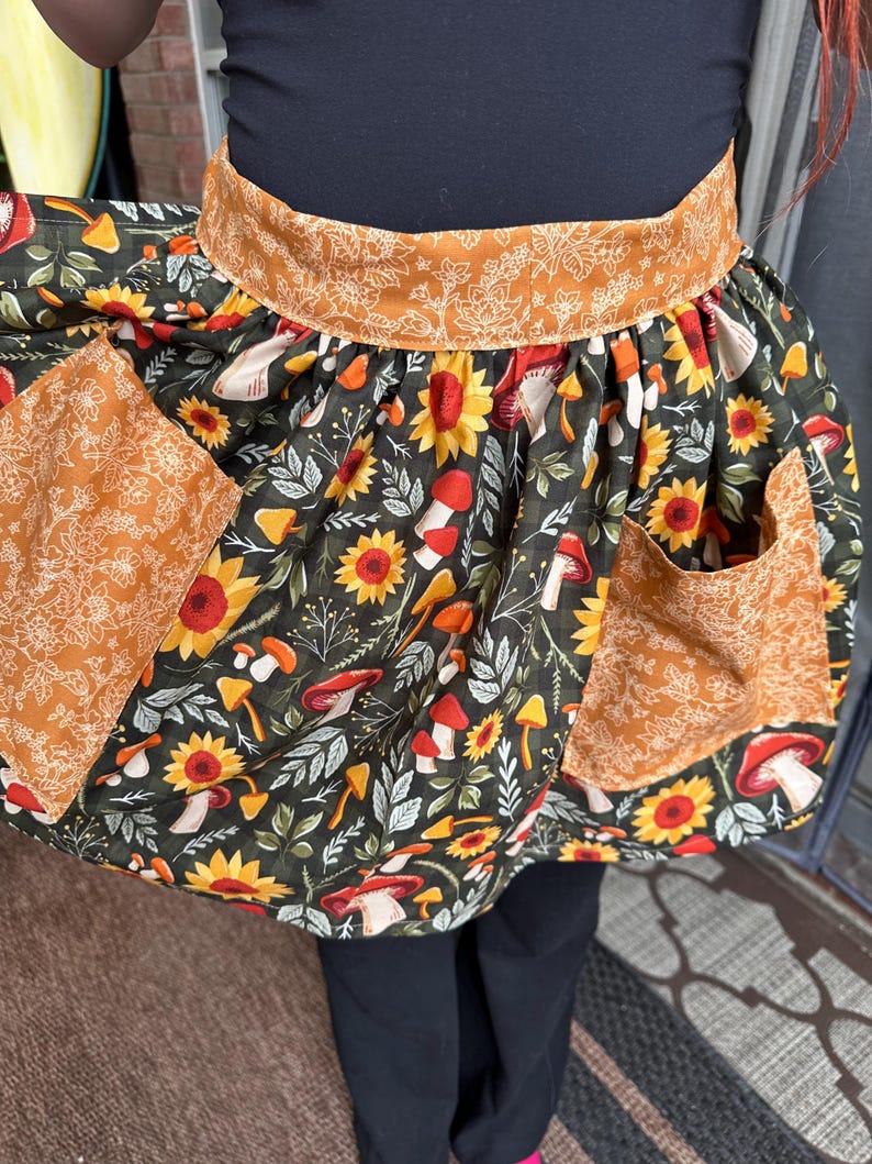K&ouml;nnte beinhalten: Ein farbenfrohe Sch&uuml;rze mit dunkelgr&uuml;nem Hintergrund, verziert mit Sonnenblumen, Pilzen und Bl&auml;ttern. Die Sch&uuml;rze hat zwei Taschen und einen Bund mit kontrastierendem orangefarbenem Blumenmuster. F&uuml;r K&uuml;che oder Bastelarbeiten.
