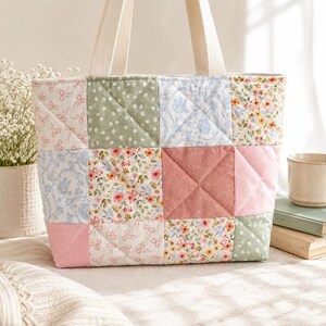 Puede incluir: Un bolso tote acolchado con un diseño de patchwork. El bolso presenta cuadrados de tela con varios estampados, incluyendo flores, lunares y lazos, en tonos rosa, verde y azul. Tiene asas color crema y una textura acolchada.