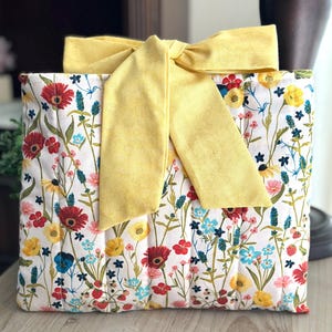 May include: A floral-patterned gift bag with a large yellow bow. The bag features a white background with a colorful array of flowers in red, yellow, blue, and pink. The bow is a solid yellow color.