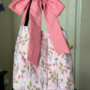 May include: A pink tote bag with a large pink bow. The bag features a strawberry and floral pattern with green leaves, white flowers, and blue bows. The bag is designed with a pleated style.