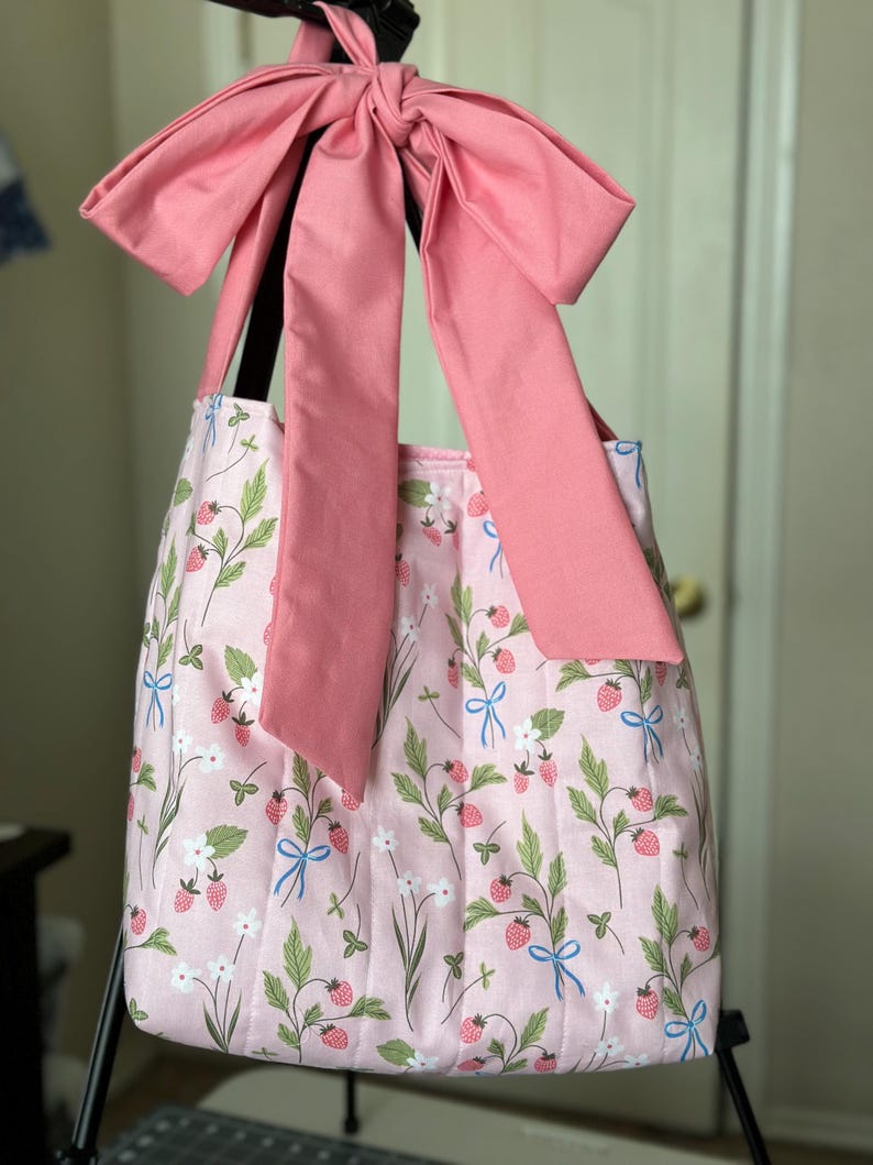 May include: A pink tote bag with a large pink bow. The bag features a strawberry and floral pattern with green leaves, white flowers, and blue bows. The bag is designed with a pleated style.