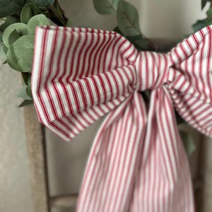 May include: A close-up of a decorative bow made of red and white striped fabric. The bow is tied and attached to a green leafy wreath. The fabric has a classic ticking stripe pattern.