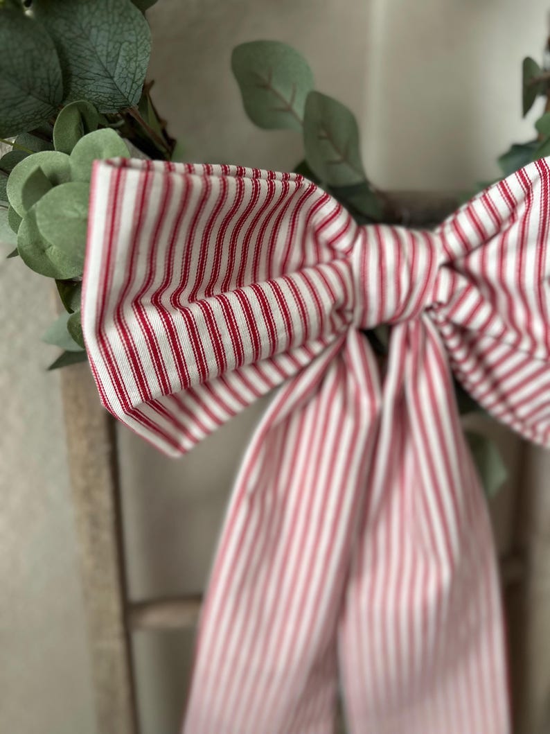 May include: A close-up of a decorative bow made of red and white striped fabric. The bow is tied and attached to a green leafy wreath. The fabric has a classic ticking stripe pattern.