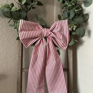 May include: A decorative wreath featuring a large bow with red and white stripes. The bow is centered on the wreath, which is adorned with green leaves and small white flowers. The wreath is hanging on a wooden ladder.