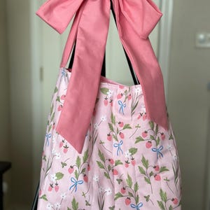 May include: A pink tote bag with a strawberry and floral pattern. The bag has a large pink bow at the top and long pink straps. The bag's fabric features green leaves, white flowers, and red strawberries with blue bows.