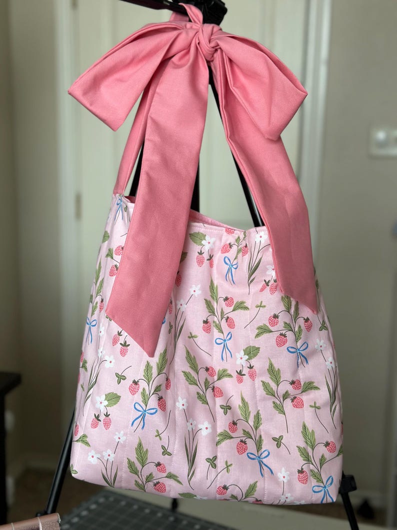 May include: A pink tote bag with a strawberry and floral pattern. The bag has a large pink bow at the top and long pink straps. The bag's fabric features green leaves, white flowers, and red strawberries with blue bows.