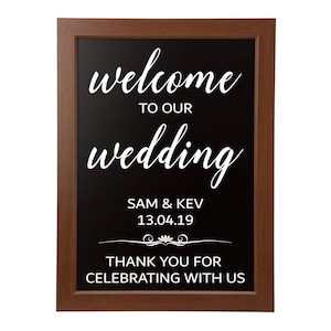 Puede incluir: Un letrero enmarcado con un fondo negro y una escritura blanca que dice "welcome to our wedding". Debajo de la escritura están los nombres "SAM & KEV" y la fecha "13.04.19". La parte inferior del letrero dice "THANK YOU FOR CELEBRATING WITH US". El marco es marrón.