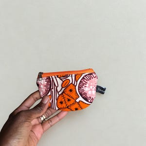 Op de afbeelding: Een kleine, bedrukte portemonnee met een oranje rits. De portemonnee heeft een levendig ontwerp met oranje, roze en witte vormen. Een klein label met het woord "Trama" is aan de zijkant bevestigd. De portemonnee wordt in een hand gehouden.