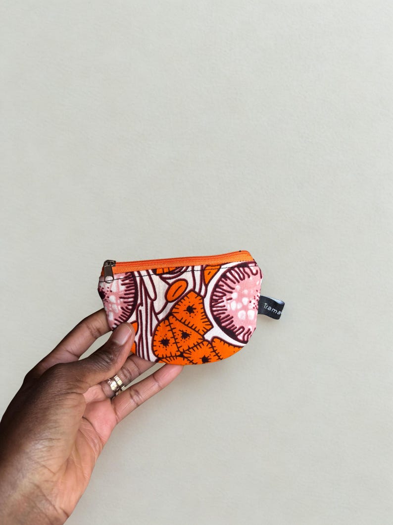 Op de afbeelding: Een kleine, bedrukte portemonnee met een oranje rits. De portemonnee heeft een levendig ontwerp met oranje, roze en witte vormen. Een klein label met het woord "Trama" is aan de zijkant bevestigd. De portemonnee wordt in een hand gehouden.