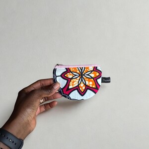 Op de afbeelding: Een kleine, halve cirkelvormige portemonnee met een levendig bloemmotief in oranje, roze en wit op een marineblauwe achtergrond. De portemonnee heeft een roze rits en een klein zwart label met "TramaC".