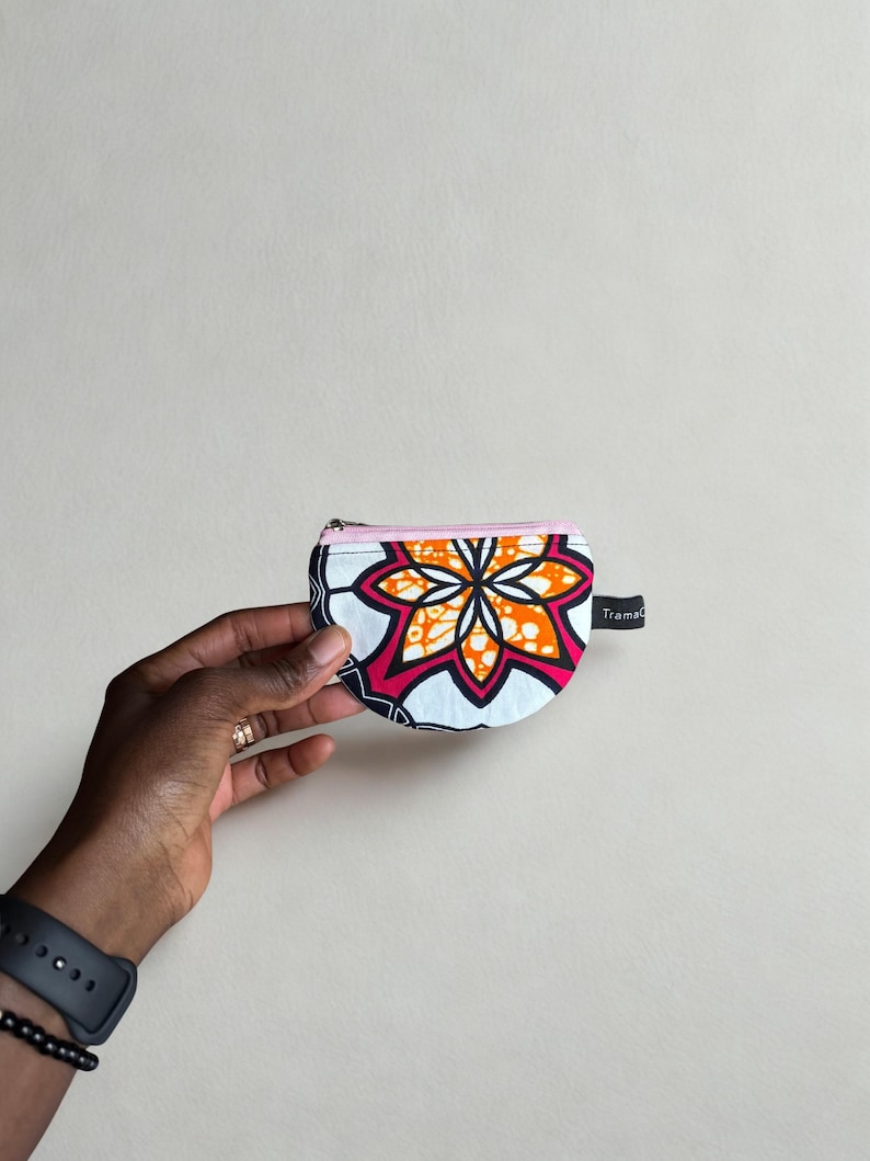 Op de afbeelding: Een kleine, halve cirkelvormige portemonnee met een levendig bloemmotief in oranje, roze en wit op een marineblauwe achtergrond. De portemonnee heeft een roze rits en een klein zwart label met "TramaC".