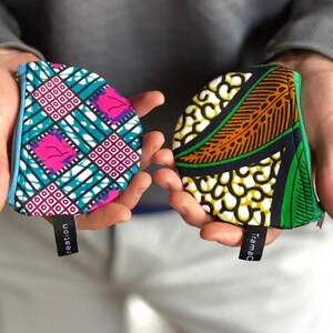 Op de afbeelding: Twee kleine, kleurrijke stoffen zakjes met ritssluitingen worden in open handen gehouden. E&eacute;n zakje heeft een turquoise, roze en wit geometrisch patroon. De andere heeft een geel, oranje en groen ontwerp. Elk heeft een klein zwart label.