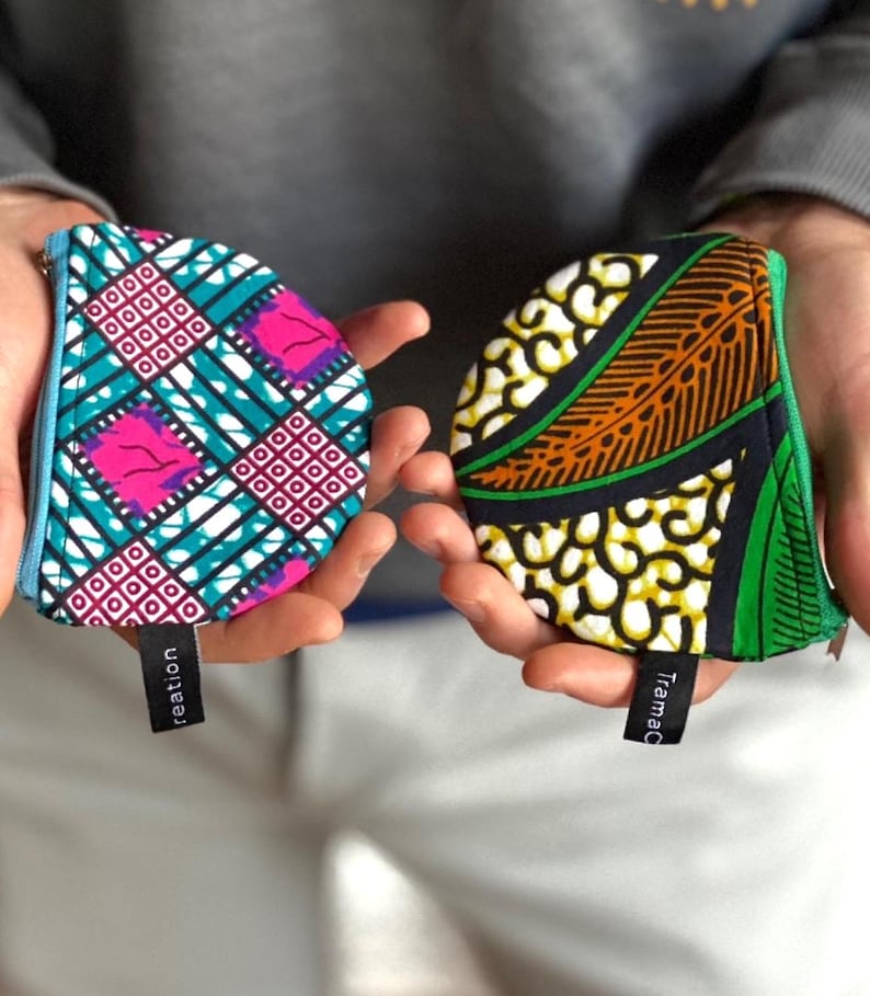 Op de afbeelding: Twee kleine, kleurrijke stoffen zakjes met ritssluitingen worden in open handen gehouden. E&eacute;n zakje heeft een turquoise, roze en wit geometrisch patroon. De andere heeft een geel, oranje en groen ontwerp. Elk heeft een klein zwart label.
