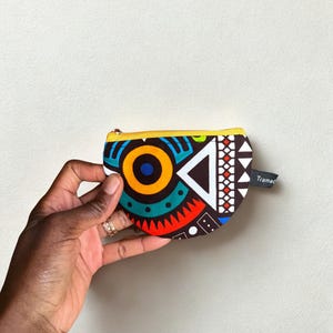Op de afbeelding: Een kleine, kleurrijke portemonnee met een geometrisch patroon in rood, blauw, geel en wit. De portemonnee heeft een gele rits en een klein label met de tekst "Tramac". De portemonnee wordt in een hand gehouden.