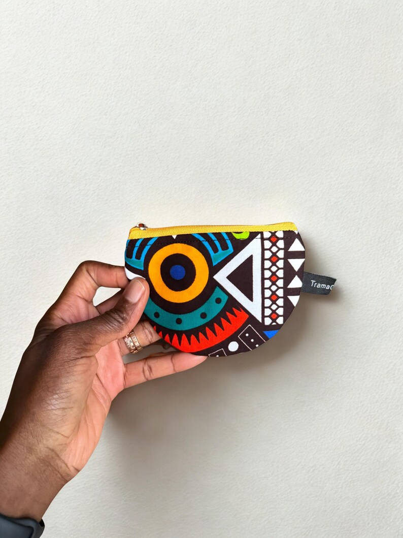 Op de afbeelding: Een kleine, kleurrijke portemonnee met een geometrisch patroon in rood, blauw, geel en wit. De portemonnee heeft een gele rits en een klein label met de tekst "Tramac". De portemonnee wordt in een hand gehouden.