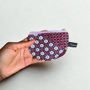 Op de afbeelding: Een kleine, bedrukte portemonnee met een paarse rits en rand. De portemonnee heeft een herhalend geometrisch ontwerp in rood, wit en blauw. De merknaam "Tramad" is gedrukt op een klein zwart label.