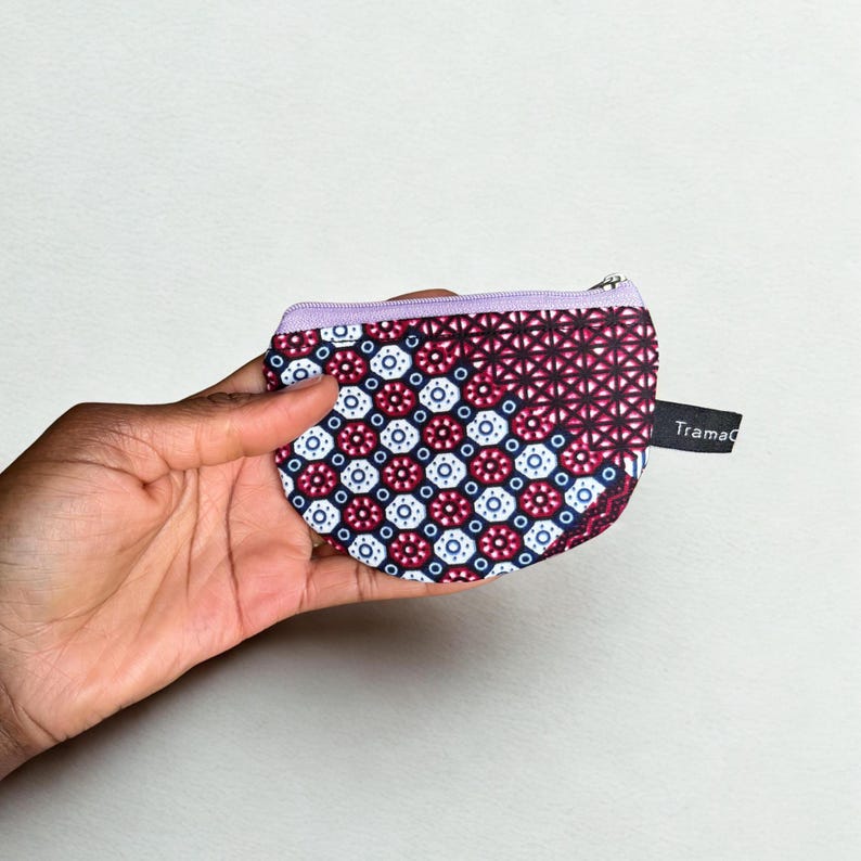Op de afbeelding: Een kleine, bedrukte portemonnee met een paarse rits en rand. De portemonnee heeft een herhalend geometrisch ontwerp in rood, wit en blauw. De merknaam "Tramad" is gedrukt op een klein zwart label.
