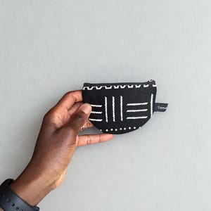 Op de afbeelding: Zwart-wit patroon muntenportemonnee met ritssluiting. De portemonnee heeft een geometrisch ontwerp met verticale en horizontale lijnen en kleine stippen. Een klein label met het woord "Trama" is aan de zijkant bevestigd.