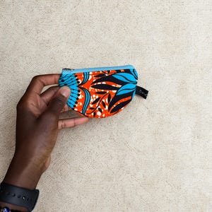 Op de afbeelding: Een kleine, kleurrijke portemonnee met een blauwe rits en een levendig patroon van oranje, zwarte en blauwe bladeren. De portemonnee wordt in een hand gehouden, wat de compacte afmetingen en het ontwerp laat zien. De tekst "Trans" is zichtbaar.