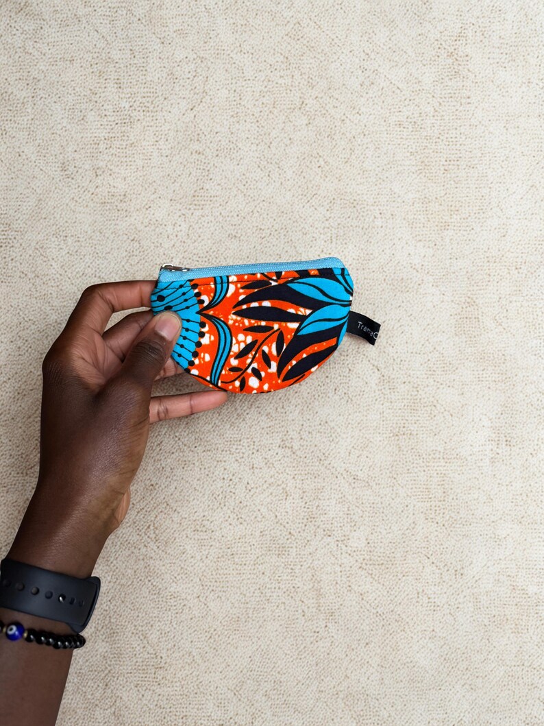 Op de afbeelding: Een kleine, kleurrijke portemonnee met een blauwe rits en een levendig patroon van oranje, zwarte en blauwe bladeren. De portemonnee wordt in een hand gehouden, wat de compacte afmetingen en het ontwerp laat zien. De tekst "Trans" is zichtbaar.