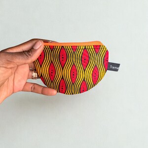 Op de afbeelding: Een kleine, ronde portemonnee met een levendige Afrikaanse print. De portemonnee heeft een herhalend patroon van rode, gele en zwarte golvende lijnen en stippen. Het heeft een oranje rits en een klein label met de tekst "Trama".