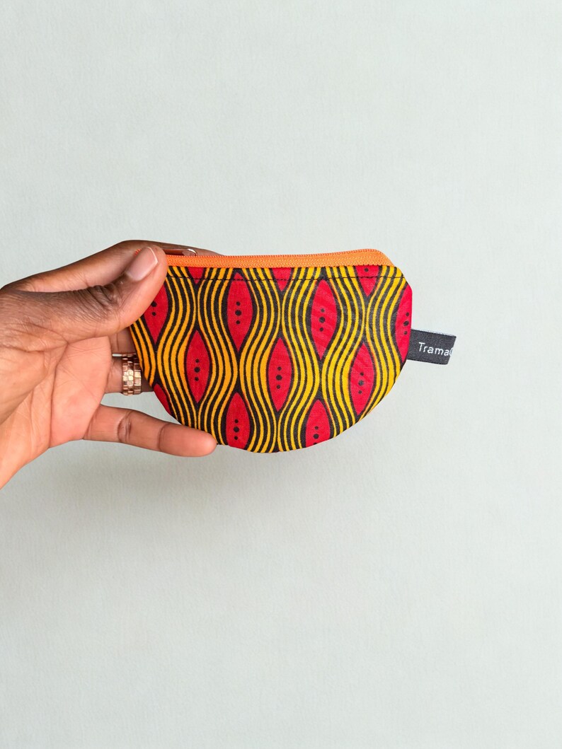 Op de afbeelding: Een kleine, ronde portemonnee met een levendige Afrikaanse print. De portemonnee heeft een herhalend patroon van rode, gele en zwarte golvende lijnen en stippen. Het heeft een oranje rits en een klein label met de tekst "Trama".