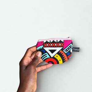 Op de afbeelding: Een kleine, kleurrijke portemonnee met een geometrisch patroon in rood, geel, blauw en zwart. De portemonnee heeft een roze rits en een klein zwart label met de tekst "TramaC". De portemonnee wordt in een hand gehouden.
