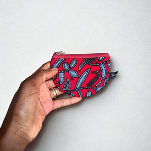 Op de afbeelding: Een kleine, rode etui met ritssluiting en een zwart en blauw bladerenpatroon. De etui wordt in een hand gehouden, waarbij de levendige kleuren en het ontwerp worden getoond. De rits is rood en de etui lijkt van stof te zijn.