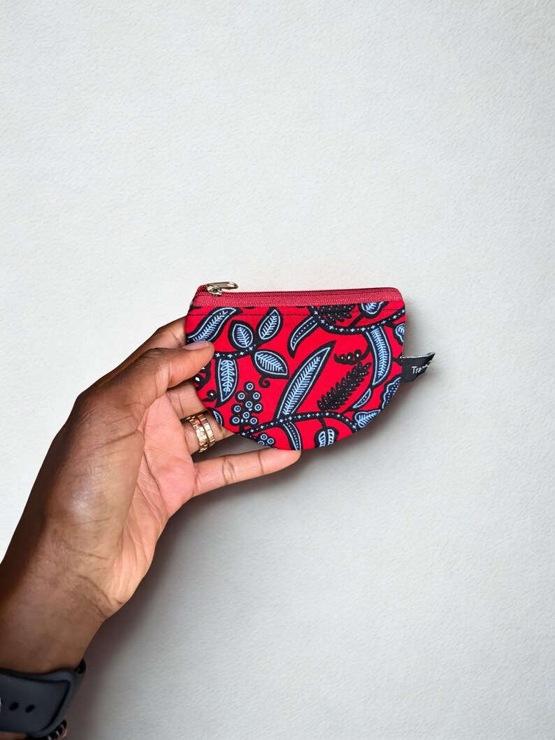Op de afbeelding: Een kleine, rode etui met ritssluiting en een zwart en blauw bladerenpatroon. De etui wordt in een hand gehouden, waarbij de levendige kleuren en het ontwerp worden getoond. De rits is rood en de etui lijkt van stof te zijn.