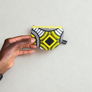 Op de afbeelding: Een kleine, bedrukte portemonnee met een gele rits en een zwart-wit geometrisch ontwerp. De portemonnee heeft een zwarte ruit in het midden en het woord "rama" op een klein label.