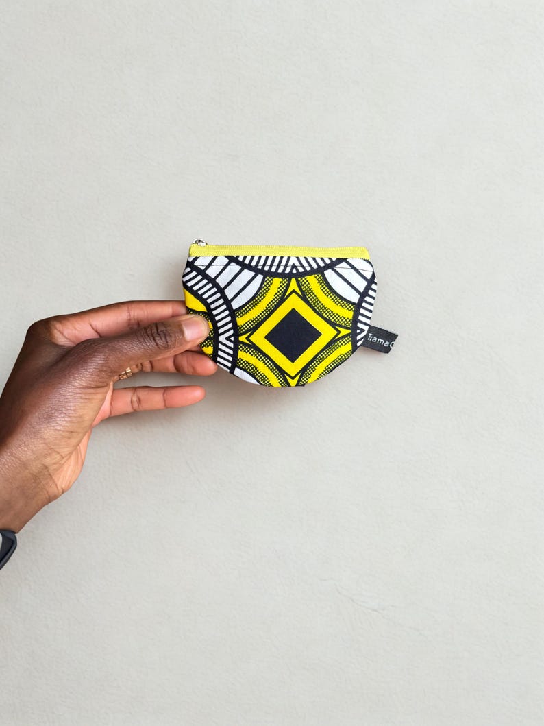 Op de afbeelding: Een kleine, bedrukte portemonnee met een gele rits en een zwart-wit geometrisch ontwerp. De portemonnee heeft een zwarte ruit in het midden en het woord "rama" op een klein label.