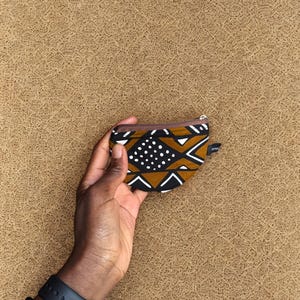 Op de afbeelding: Een kleine portemonnee met een bruine rits, met een geometrisch patroon in zwart, wit en bruin, wordt tegen een getextureerde bruine achtergrond gehouden. Een zwart horloge en een kralenarmband zijn zichtbaar op de pols.