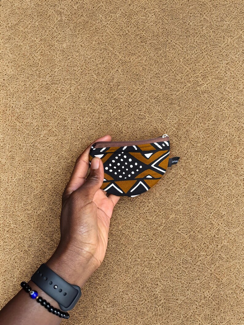 Op de afbeelding: Een kleine portemonnee met een bruine rits, met een geometrisch patroon in zwart, wit en bruin, wordt tegen een getextureerde bruine achtergrond gehouden. Een zwart horloge en een kralenarmband zijn zichtbaar op de pols.