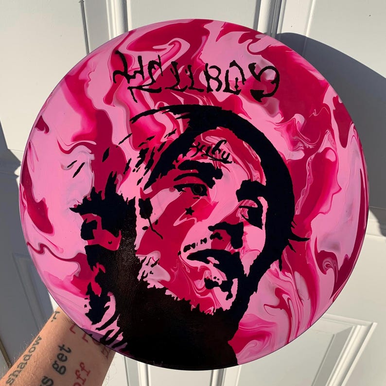 May include: A circular disc featuring a marbled pink and magenta background with a black silhouette portrait of a person. The text HELLBOY is printed in black at the top, and the word Peep is written in black script above the person's forehead.