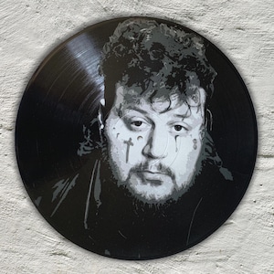 May include: A black vinyl record with a white stencil portrait of a man with a beard and tattoos. The man is looking directly at the viewer with a serious expression.