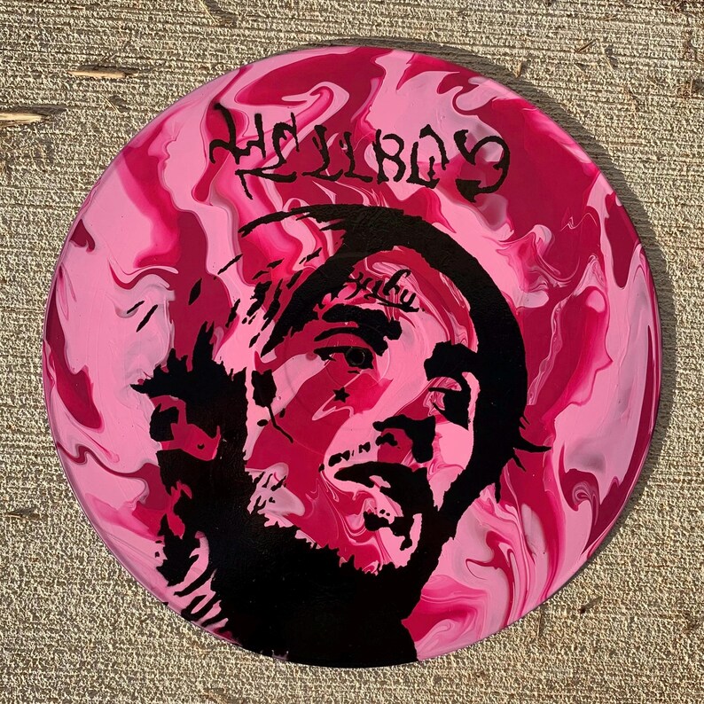 May include: A circular vinyl record featuring a pink and white marbled background with a black silhouette portrait of a person. The text HELLBOY is printed in a stylized black font at the top, and the word baby is visible on the forehead area.
