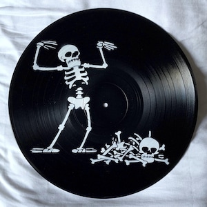 May include: A black vinyl record featuring a white skeleton design. The skeleton is depicted with arms raised, and a pile of bones and a skull are at the bottom. The record has grooves and a central label area.