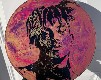 May include: Round artwork featuring a portrait of a person with black hair and a black silhouette. The background has a colorful, swirled design in shades of pink, purple, and gold. The artwork is held by a person wearing a black sleeve.