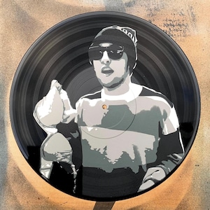 May include: A black vinyl record with a white and gray stencil of a person wearing a black beanie and sunglasses. The person is looking directly at the viewer and has a serious expression.