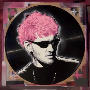 May include: A black vinyl record featuring a stencil portrait of a person with pink hair and sunglasses. The artwork is in shades of black, white, and pink, with a textured background and pink cross-shaped accents.