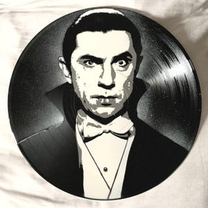 May include: A black vinyl record with a white stencil of a man's face. The man is wearing a tuxedo and has a serious expression. The record is a popular art medium for creating unique and personalized pieces.