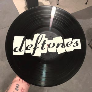 May include: A black vinyl record with the band name "Deftones" in white, stylized lettering. The record is circular with grooves visible on the surface. The image is taken from a slightly elevated angle.