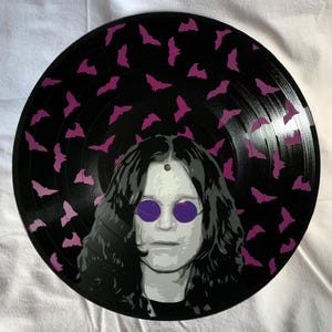 May include: A black vinyl record with a purple and black design of bats and a portrait of a person with long dark hair wearing purple sunglasses.