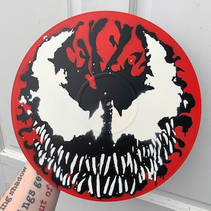 May include: A red vinyl record with a black and white spray paint design of a Venom character. The design features a black and white silhouette of Venom's head with a red background.