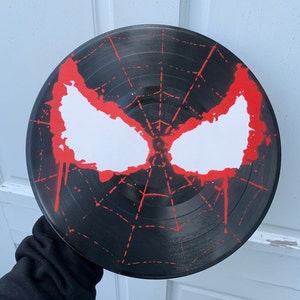 May include: A black vinyl record painted with a red and white spider-man design. The design features a web pattern and a white spider-man mask with red dripping paint.