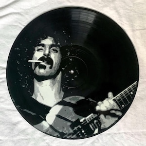 May include: A black vinyl record featuring a painted portrait of a musician with a mustache, smoking a cigarette, and playing a guitar. The artwork is in shades of gray and white against the black record.