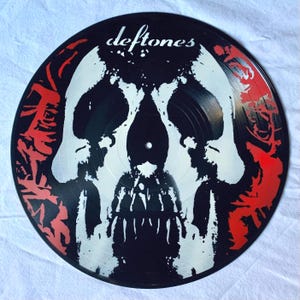 May include: Black vinyl record with a white skull graphic and the band name "Deftones" printed in white at the top. The skull has a red and black background.