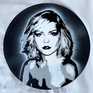 May include: Black and white spray paint portrait of a woman with short, choppy hair on a black vinyl record.