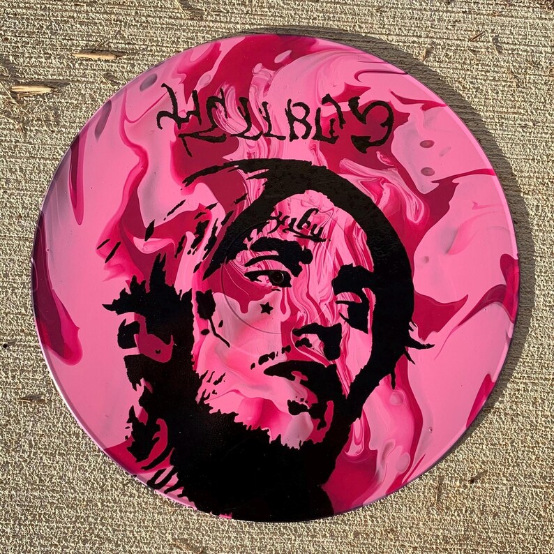 May include: A circular disc featuring a marbled pink and magenta background with a black silhouette portrait of a person with facial tattoos. The text HELLBOY is printed in a stylized black font at the top, and the word BABY is visible on the forehead.